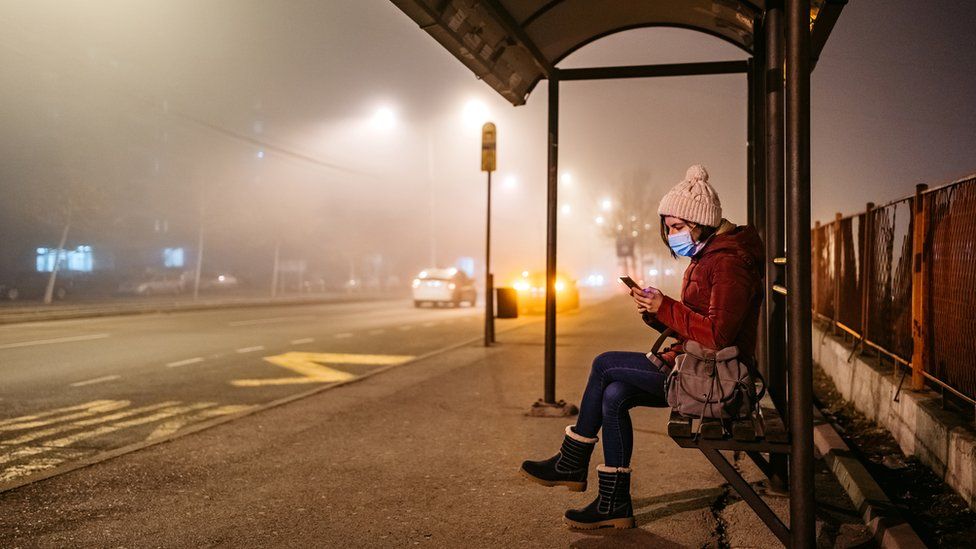 woman wearing mask waits at a bus stop on a foggy night