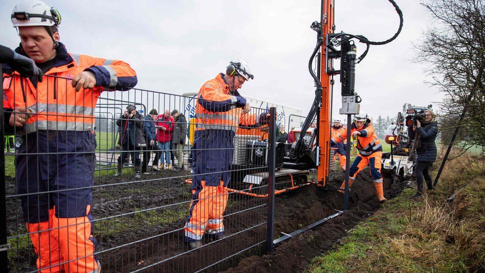 Workers in orange uniforms assemble the first portions of a metal fence on the Danish border, while camera crews look on nearby