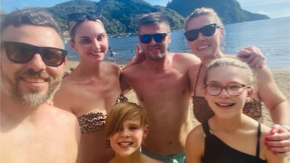 Four adults and two children posing for a selfie on a beach. The sun is shining and they all look happy.
