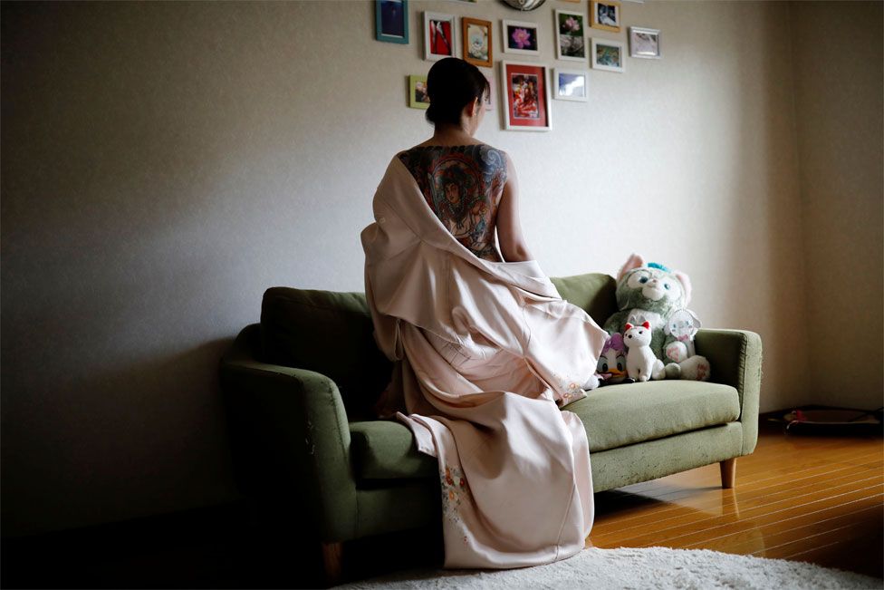 A woman kneels on a couch with a garment pulled down to reveal her large back tattoo to the camera