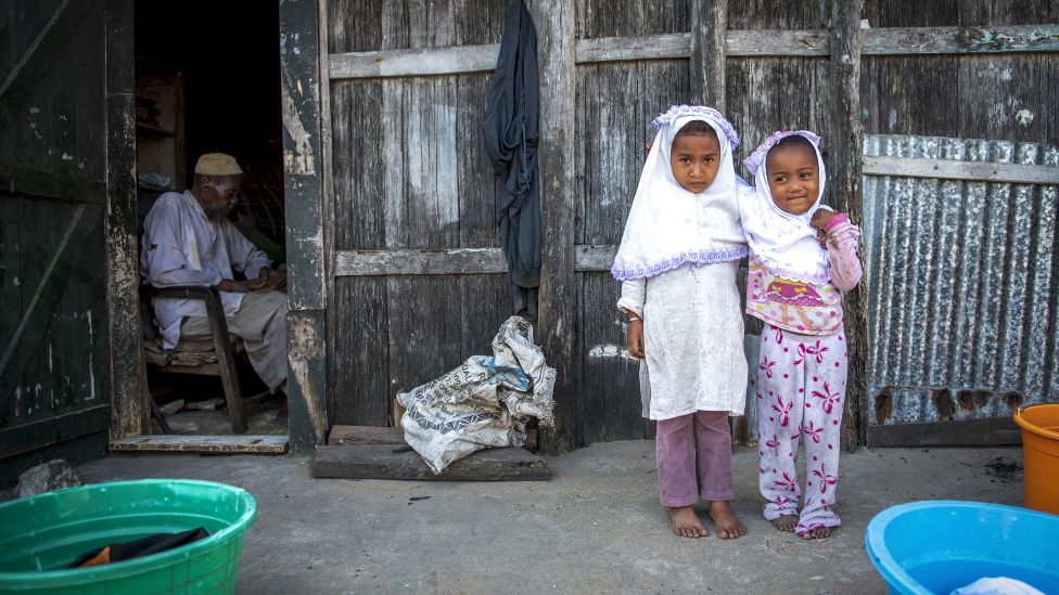 Two girls pose for a photograph as an old man read Quran in his home in Fort Dauphin, Madagascar