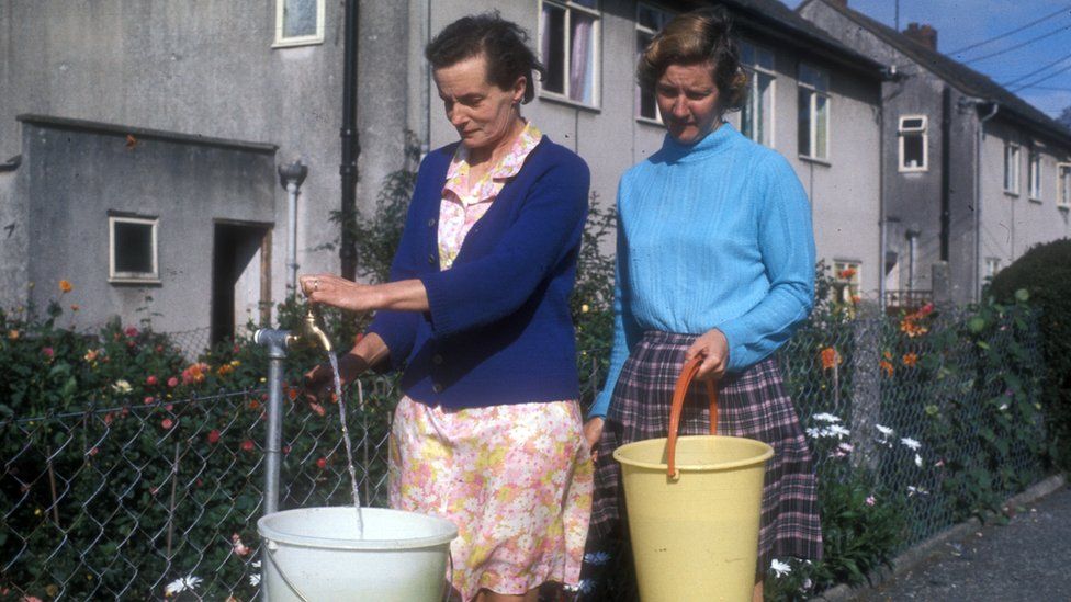 Two women holding buckets to get water from standpipes on the street