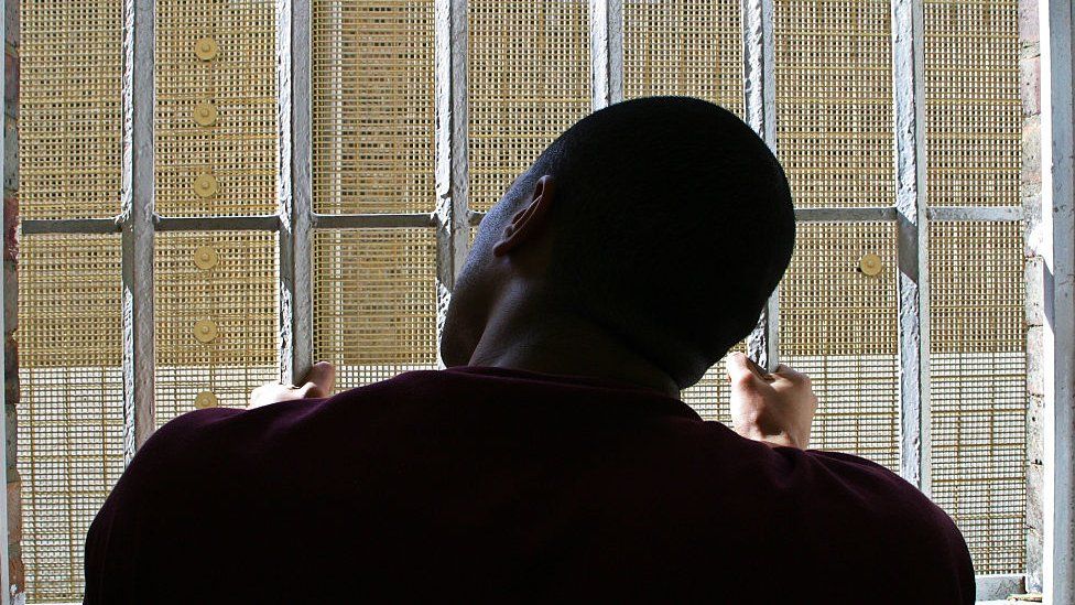 Молодой чернокожий заключенный смотрит через решетку окна своей камеры в крыле D тюрьмы Уондсворт в Лондоне. HMP Wandsworth на юго-западе Лондона была построена в 1851 году и является одной из крупнейших тюрем в Западной Европе. Он рассчитан на 1456 заключенных.