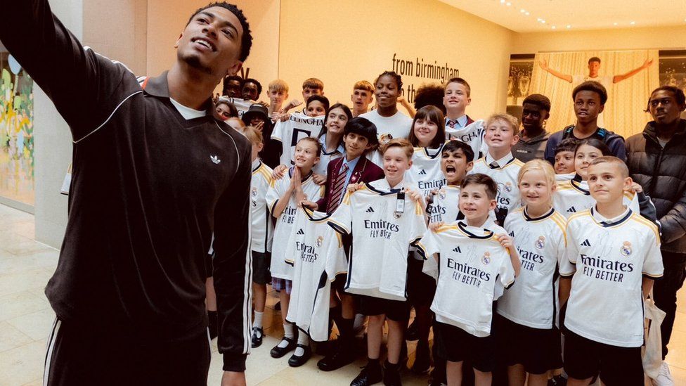 Jude Bellingham holds a phone and takes a selfie, behind him is a group of school children, holding Real Madrid shirts.