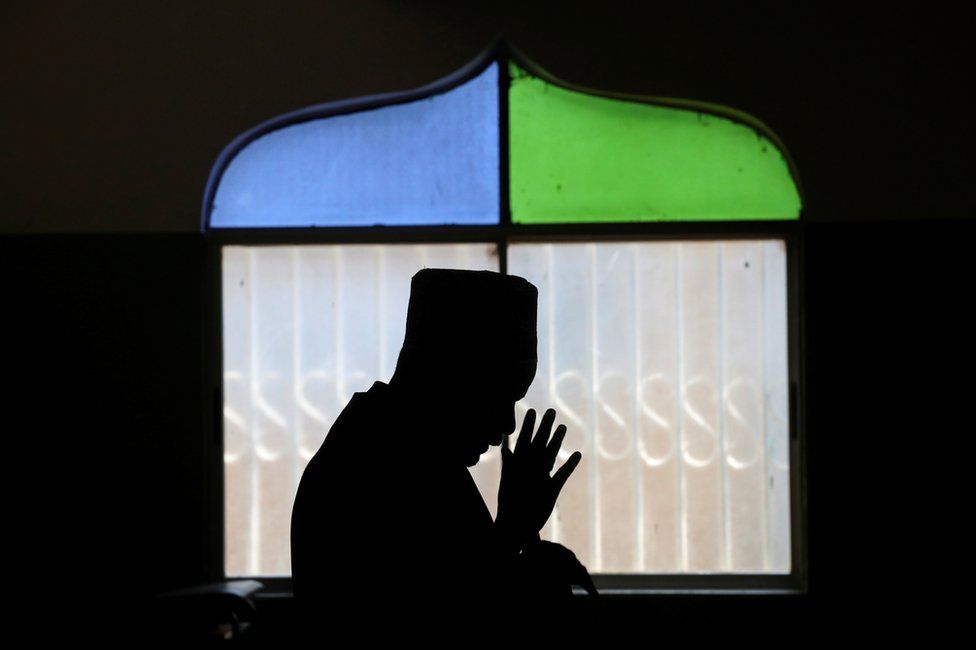 A man prays in a mosque. His silhouette is seen against the window.