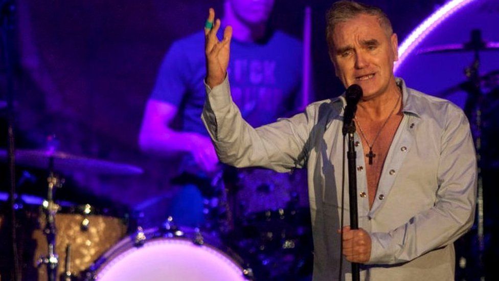 A recent photo of Morrissey during a performance. He is standing at a microphone and singing.