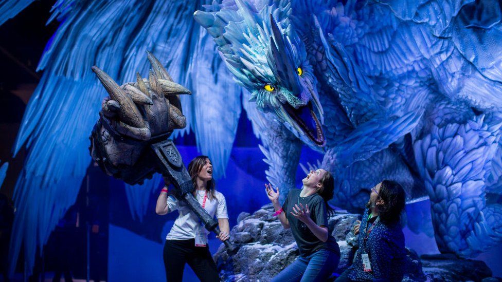 Three people cower beneath a gigantic scultpure of a dragon, with one of the people wielding a comically oversized fantasy hammer