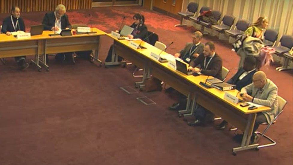 Meeting of councillors sitting behind desks. One has left her seat and is heading out of frame carrying her bag and coat.
