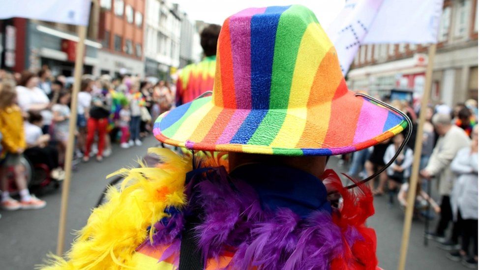 A marcher stands in front of the camera with their back to the photographer. They are wearing a rainbow trilby hat and a rainbow feather boa.