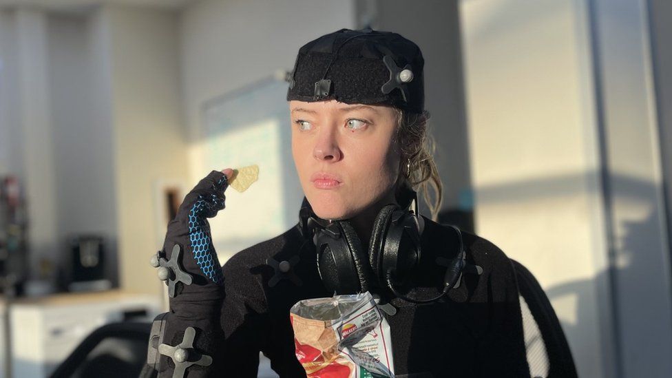 A woman sitting in a work canteen/kitchen and eating crisps while wearing a motion capture suit. It's skin tight, with small, ping-pong style balls dotted around the surface. She's also wearing large, over-ear headphones around her neck.