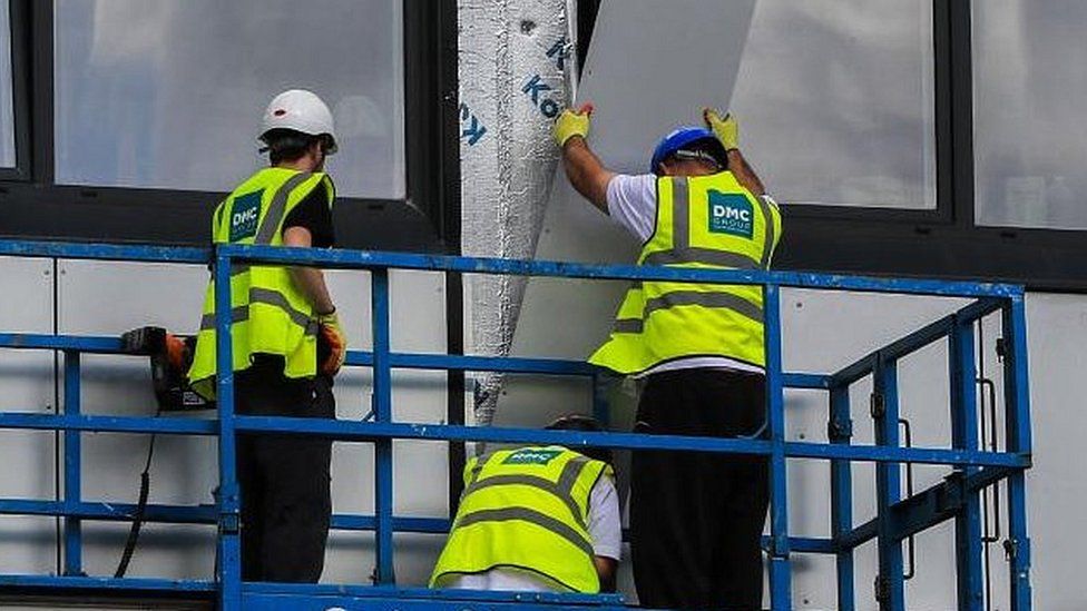 Three men in yellow hi-viz remove a panel from a block of flats