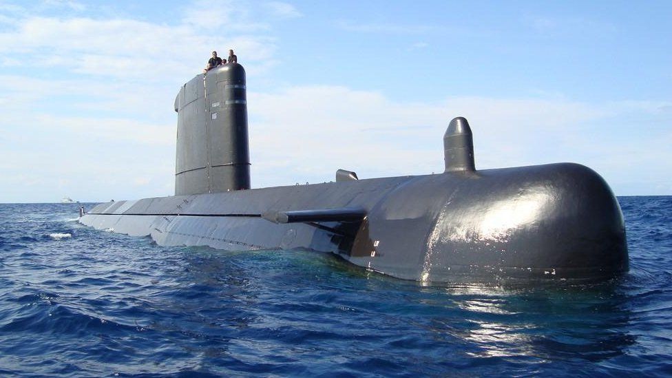 A submarine floats on the water's surface, as two people can be seen sitting on its vertical fin