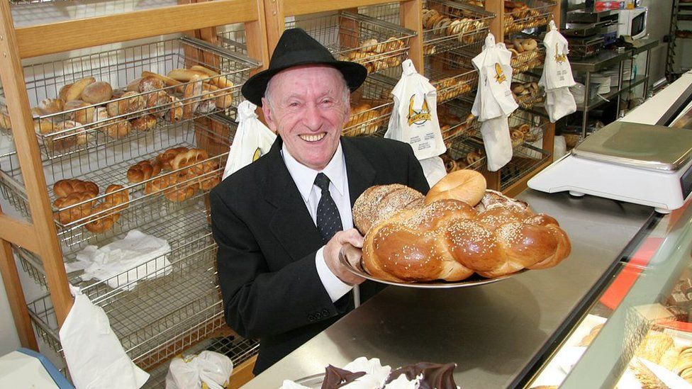 Mendel Glick holds a plate of bagels and other baked items behind the counter at Glick's