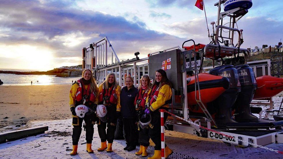 Five women stand next to a lifeboat