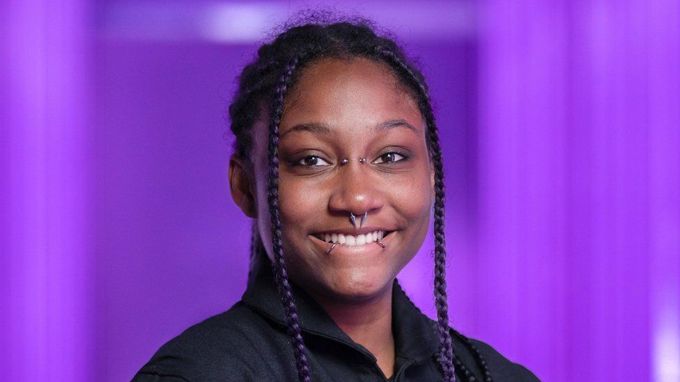 Anastatia Mayers pictured before her voyage to space. She is a young black woman with her hair worn in long braids, some coloured white and purple, tied back in a pony tail. She has piercings on the bridge of her nose, her septum and her lip and is smiling at the camera. She wears a black space suit and is photographed in front of a bright purple backdrop.