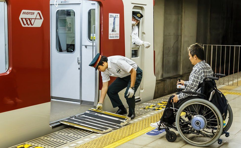 A wheelchair user boarding a train in Japan. A train worker is putting the ramp out for him.