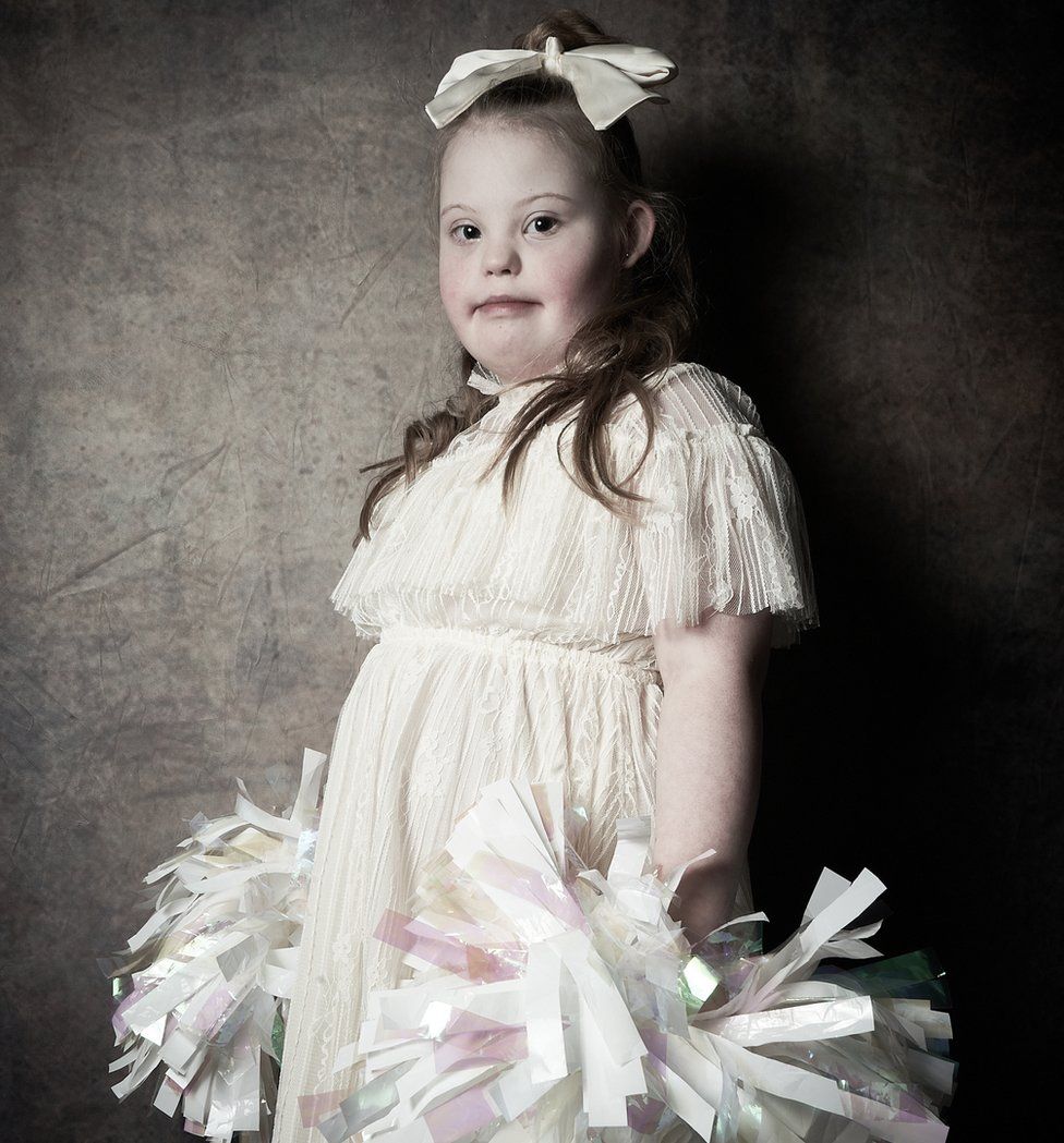 Maisie, aged eight, wearing a white dress and holding two large pom poms