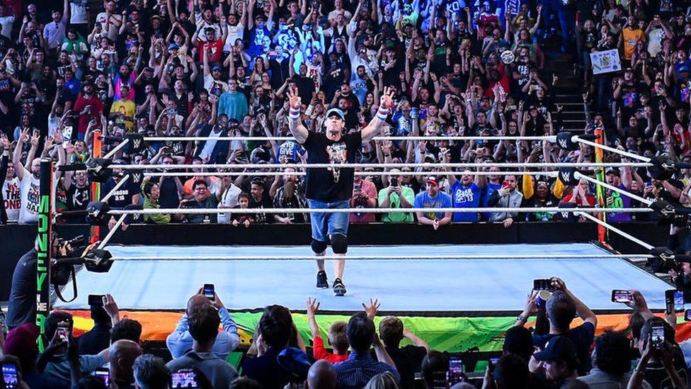 John Cena in the wrestling ring at the O2 in London. He is wearing a black t-shirt and a light blue cap and has red, white and blue sweat bands on both wrists. He's also wearing denim shorts and has black knee pads on. He has both arms raised to the sky and the crowd behind him all have their hands in the air and are cheering.