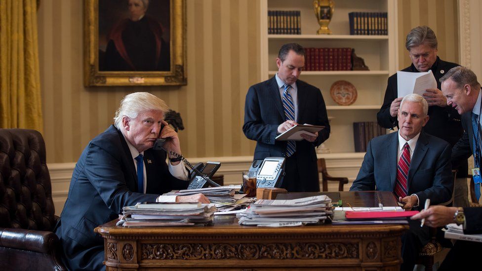 Donald Trump, seated at the desk of the Oval Office, speaks on the phone, surrounded by his chief of staff Reince Preibus, VP Mike Pence, strategist Steve Bannon, and press secretary Sean Spicer