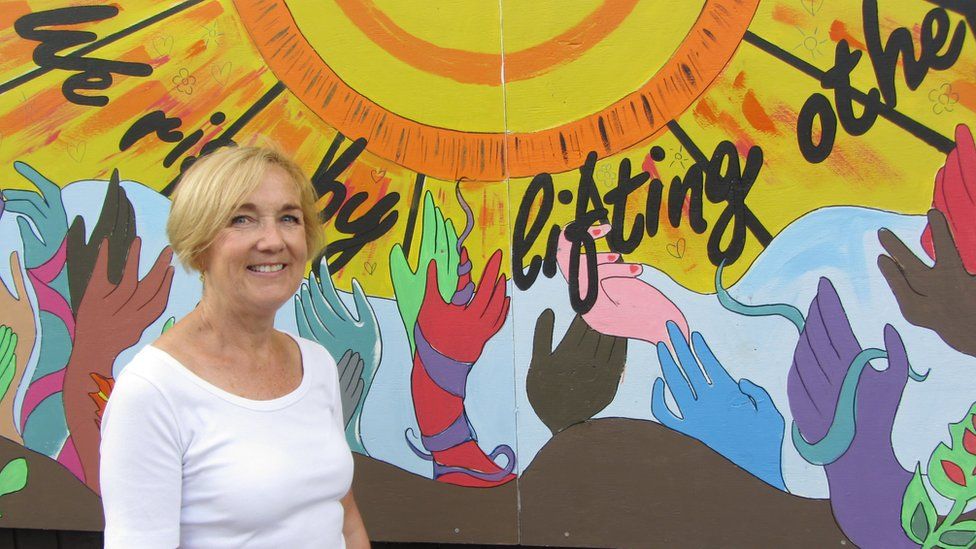 Claire Garrett, CEO of The Harbour Project standing in front of the mural with the works "we rise by lifting others" behind her