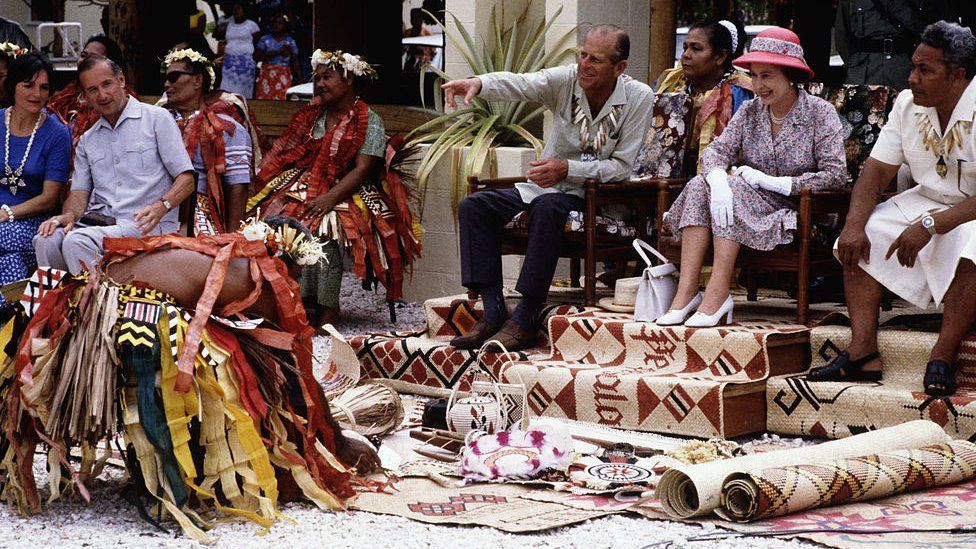 Queen Elizabeth II and Prince Philip, Duke of Edinburgh receiving gifts, while watching and photographing traditional dancers in Funafuti in Tuvalu on 27 October 1982 during the Royal Tour of the South Pacific.
