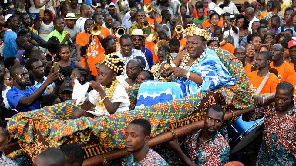 Man wearing a crown and turquoise robes carried on a litter through a large crowd