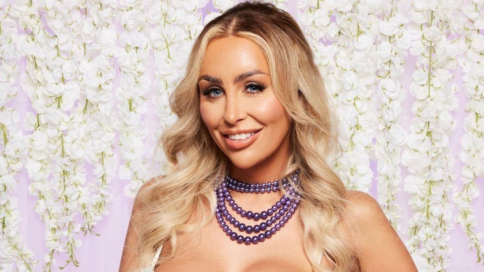 Promo shot of Ella Morgan Clark for Married at First Sight UK. Ella is a 29-year-old white woman, she has long curled blonde hair and blue eyes. She smiles at the camera, wearing a pink lip gloss and false eyelashes. She has a four-layered purple pearl necklace around her neck and the white straps of her wedding dress can be seen over her shoulders. She is photographed in front of a studio background of white wisteria flowers laid over pink.