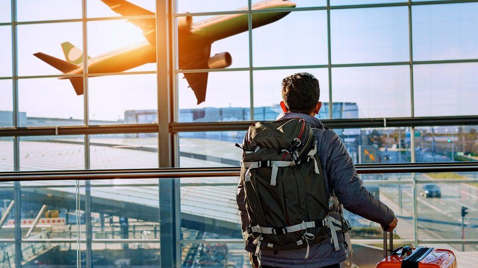 Мужчина-турист стоит в аэропорту и смотрит на полет самолета через окно.  Он держит билеты и чемодан