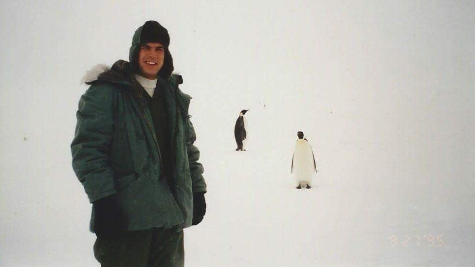 A man in winter clothing stands in front of a pure white background, with two penguins stood nearby.