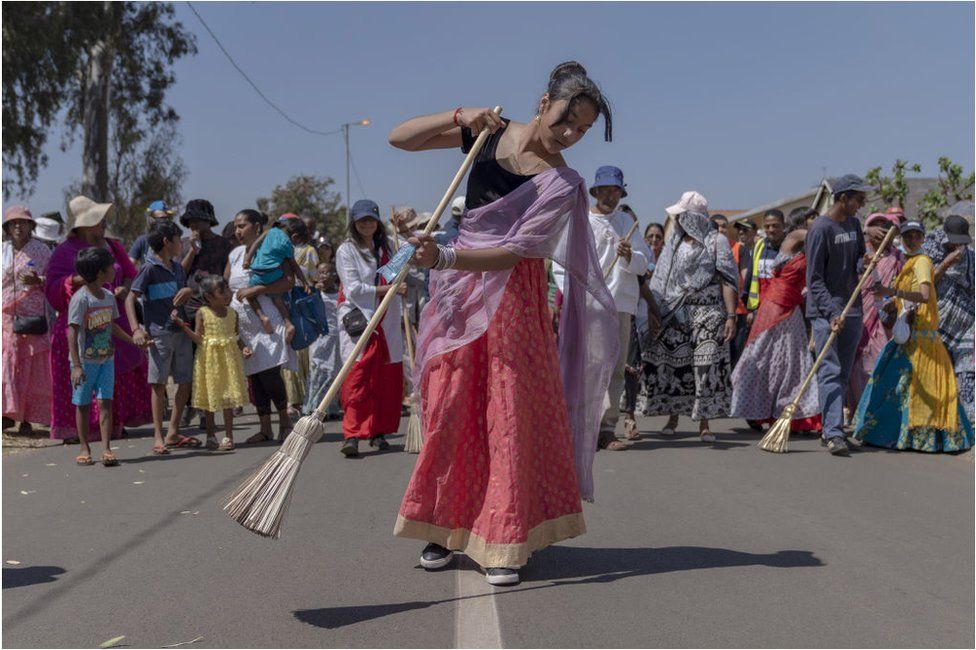 ผู้หญิงสวมชุดอินเดียโบราณกับไม้กวาด .  มีอีกหลายคนอยู่ข้างหลังเธอยืนอยู่บนถนน  มีท้องฟ้าสีคราม