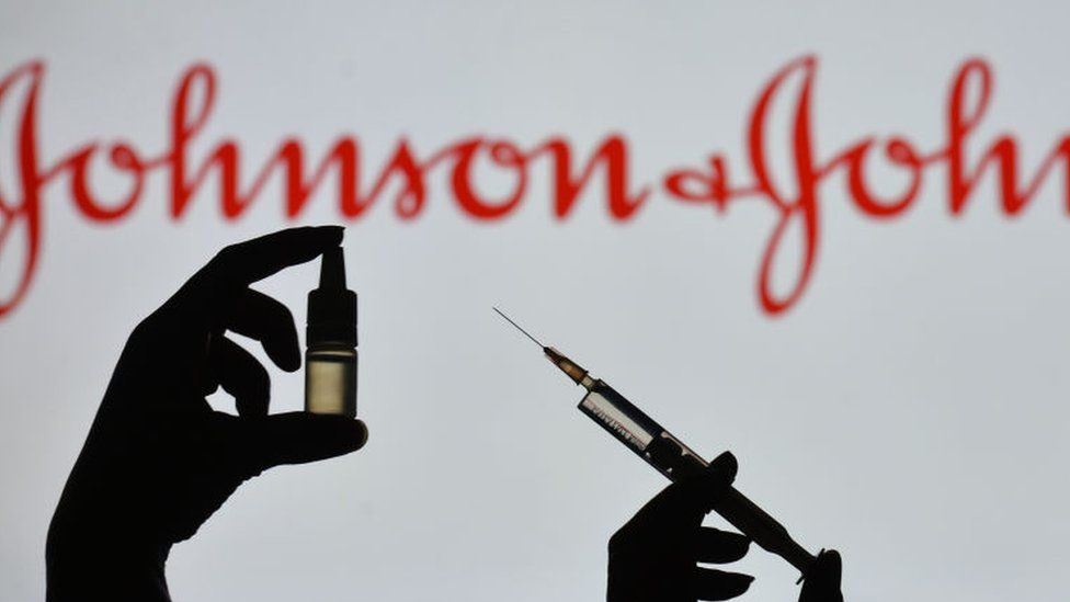 Silhouette of a man holding a medical syringe and a vial seen displayed in front of the Johnson and Johnson logo on a screen