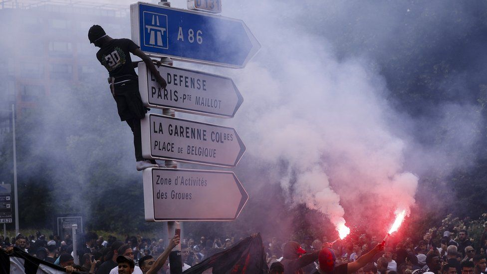 A person climbs a traffic sign as others hold flares during a march