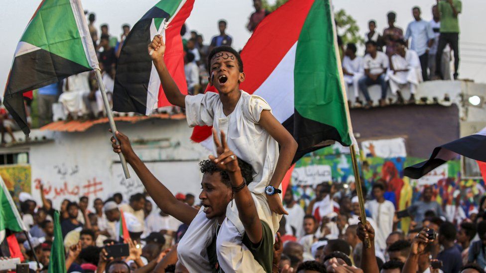 A boy carrying a Sudanese flag sits on a man's shoulders, amid a celebrating crowd