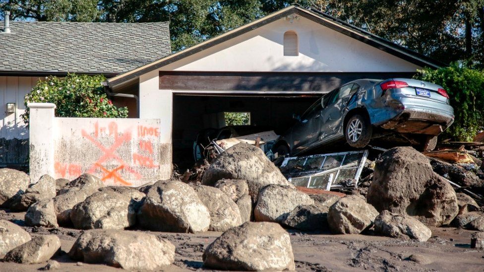 A house is shown destroyed by large boulders, with emergency service writing at the entrance and cars crushed in the drive way