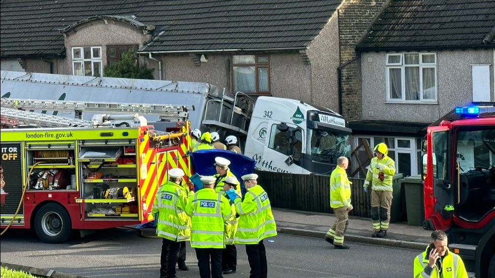 White lorry with smashed windscreen appears to be in contact with a row of houses after apparently leaving the road. Firefighters and police in hi-vis stand in the foreground on the road, with two fire engines visible