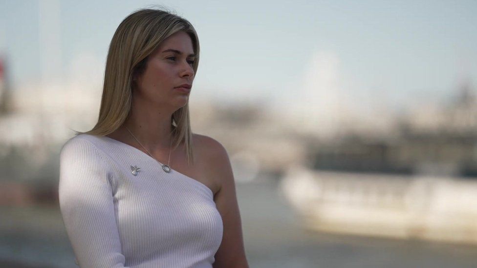 An image of Lauren, a young woman with light hair wearing a light coloured top and an angel brooch