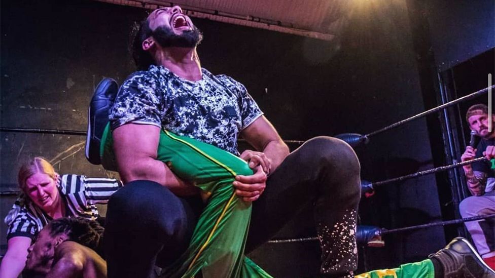 Adil Malik, wearing a printed tshirt, mid-wrestling move in a ring grappling the leg of his opponent who is wearing green wrestling tracksuits. Adil is screaming, while a female referee in the background wearing a black and white tshirt is looking at his opponent.