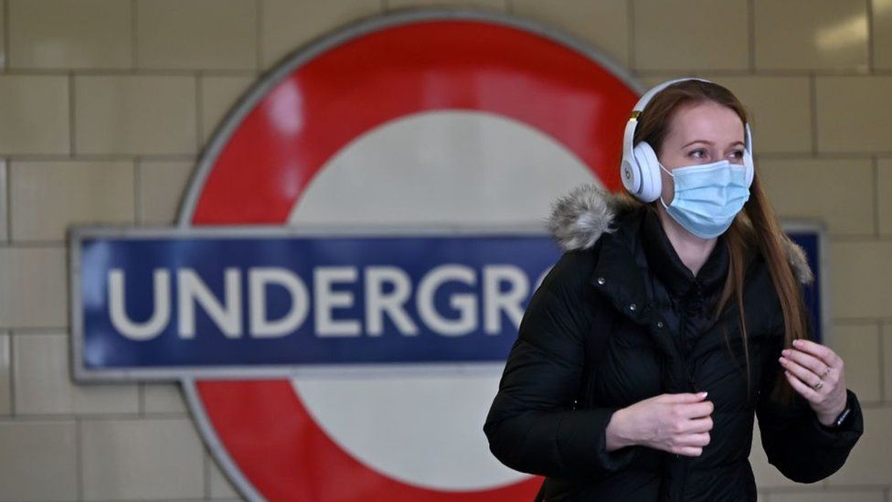 A woman wearing a mask stands in front of a London Underground sign