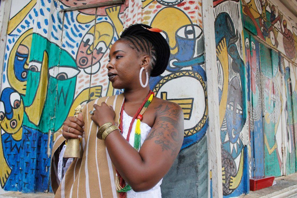 A woman stands with her side profile in front of colourful Nigerian artwork. She is wearing traditional clothing including a white outfit and braided hair. She also has on a colourful necklace.