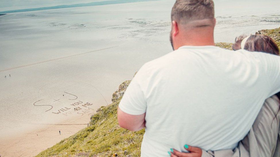 The couple look down at the proposal message written in the sand below