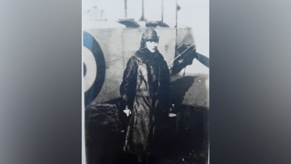 A black and white image of the female aviator wearing a long leather coat and flight head gear, standing in front of a propeller plane