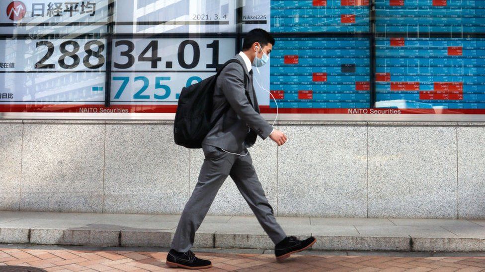 A pedestrian wearing a mask walks by an electronic board showing share prices in Tokyo.