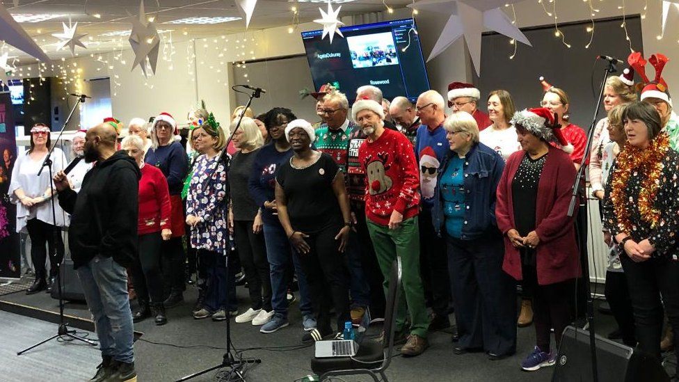 Group of people standing in a line wearing Christmas decorations. A man is talking into a microphone.