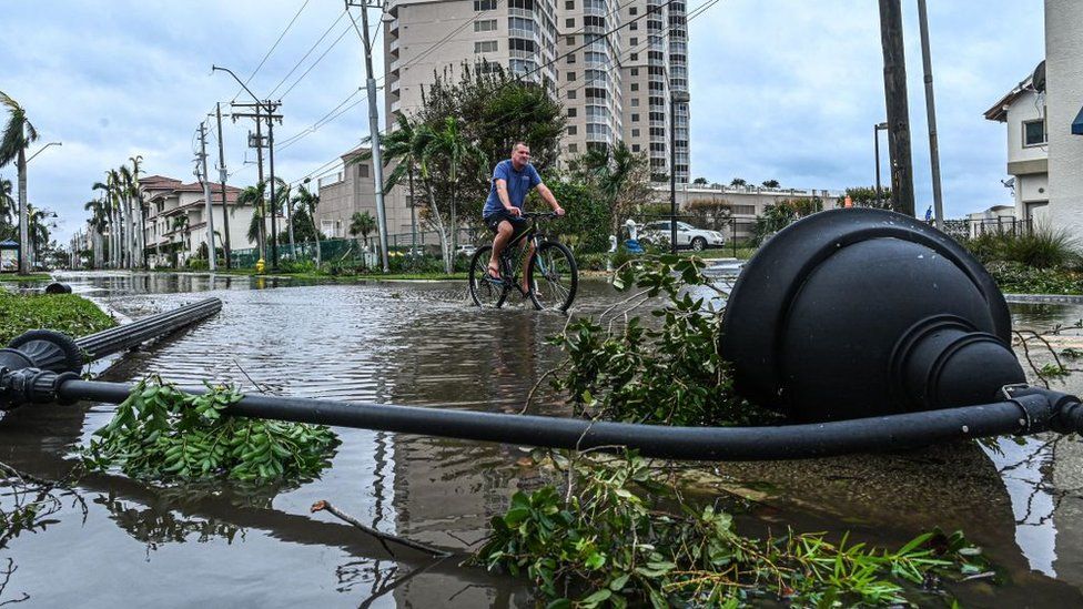 A man cycles through water past a downed street lamp in the aftermath of Hurricane Ian in Fort Myers, Florida, on September 29, 2022