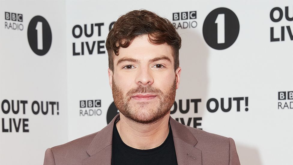 Jordan North pictured at a BBC Radio 1 event. Jordan is a white man in his 30s with short curly brown hair and a short brown heard. He has brown eyes and looks at the camera, wearing a brown suit jacket over a black top. Behind him is white and black Radio 1 branded event hoarding