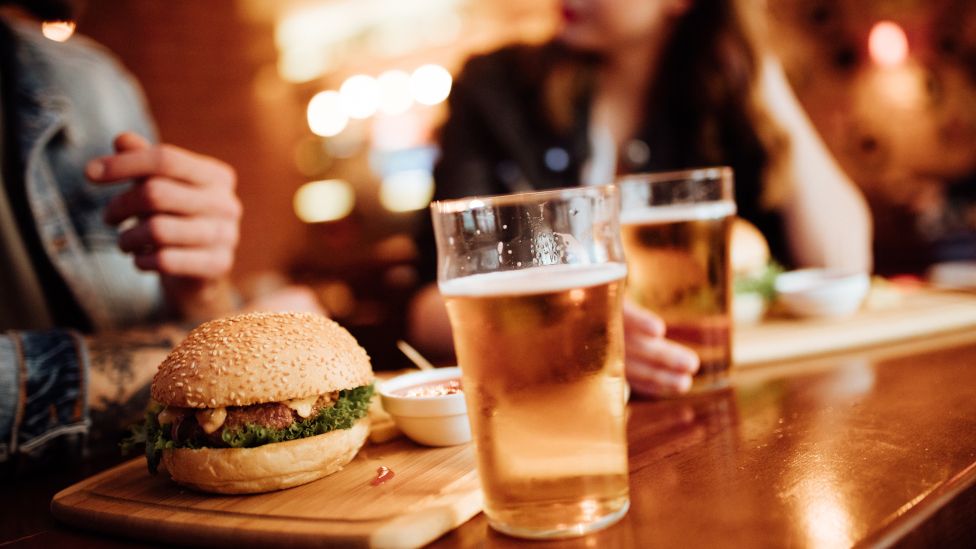 Burger, and pint glasses, with man and woman in background inside a pub