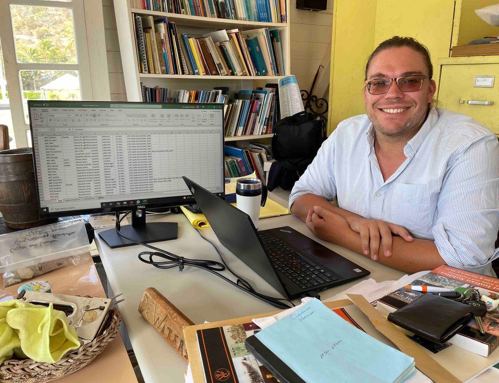 Dr Christopher Waters smiles at his desk, with a laptop and a separate desktop computer facing him. Books and files are visible on the desk and in the background.