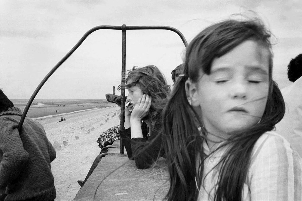 A young girl with eyes closed in front of a person looking at the sea, in Towyn