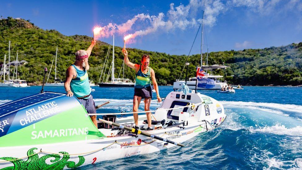 Simon and Nina Crouchman stand up in boat holding flares to celebrate arriving in Antigua after completing Atlantic row challenge