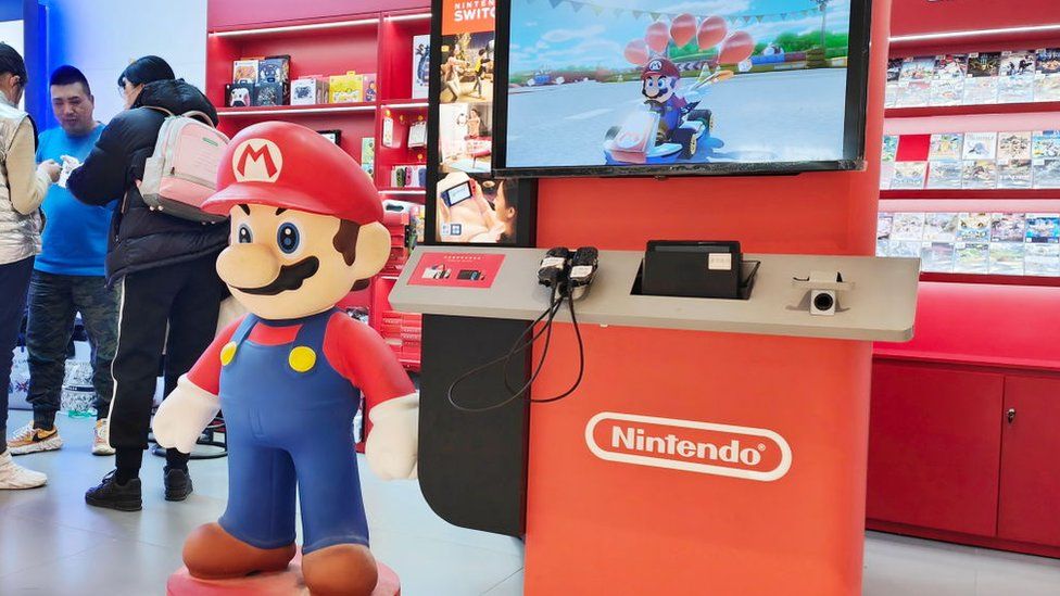 A plastic statue of Mario - complete with blue dungarees and red plumber's hat with a letter M on the brim, stands next to a Nintendo Switch demo unit. It's a large screen on a plinth with two controllers attached. On the screen is footage of Mario in a go-kart from Mario Kart. In the background are red shelves stocked with rows of Nintendo Switch games and accessories.
